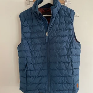 Blå dunväst från Dressmann - Säljer en blå quiltad dunväst från Dressmann med dragkedja framtill och två fickor. Västen har en hög krage och en liten brun logodetalj nertill. Perfekt för lager-på-lager och enkel att matcha med hoodies eller tröjor. Skick 9/10