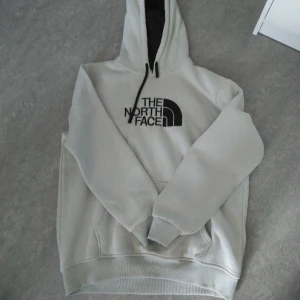 Vit& grå hoodie från The North Face - Snygg vit& grå hoodie från The North Face med svart logga på bröstet. Tröjan har huva med svarta snören, känguruficka och ribbade muddar. Perfekt för dig som gillar en clean och stilren look. Passar dig som vill ha en bekväm och trendig tröja.