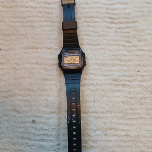 Casio F-105 digital klocka svart - Klassisk digital klocka från Casio, modell F-105. Svart plastarmband och fyrkantig display med blå detaljer. Klockan har alarm, kronograf, belysning och är vattentålig. Baksidan är i rostfritt stål. Perfekt för dig som gillar retrostil och funktion. OBS! Har en en miss färgning på bandet men syns knappt. Passar barn från 7-13 år.