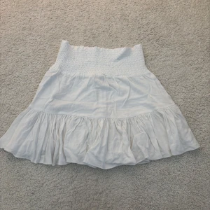 Vit volangkjol med smock - Supersöt vit kortkjol med bred smock i midjan och volang nertill. Storlek S från lager 157.