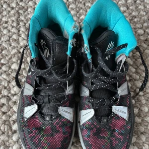 Nike Kyrie basket sneakers svart/turkos - Färgstarka Nike Kyrie 7 Pixel camo basket sneakers med svart mesh, turkos foder och coolt mönster i rosa, grönt och grått. Snygga detaljer med silverfärgade partier och svart/vita snören. Sula i grått med rosa logga undertill. Perfekta för dig som gillar att sticka ut på planen. Ordinarie pris: 2300kr (ingen originalkartong medföljer)