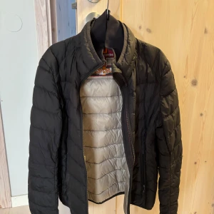 Svart lättviktsdunjacka Parajumpers Y-M - Svart quiltad lättviktsdunjacka från Parajumpers i storlek Y-M. Jackan har dragkedja, hög krage och klassisk Parajumpers-logga på ärmen. Tillverkad i syntetmaterial och dun, perfekt för kyliga dagar. Snygg och stilren design med lång ärm.Den har ett hål på höger underarm där jag har satt en svart tyg/tejp över då det smälter in, annars kan man laga det om man är händig. I övrigt väldigt fint skick. Storlek M i barn storlek. Har inte använts på flera år utan bara hängt i garderoben.