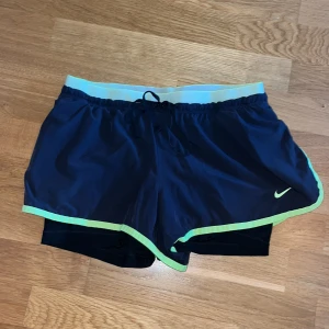 Nike Dri-Fit svarta träningsshorts - Nike Dri-Fit träningsshorts i svart med ljusgrön kant och elastisk midja. Shortsen har snörning i midjan och innerbyxa för extra komfort. Perfekta för löpning eller gym, tillverkade i lätt och snabbtorkande polyesterblandning.