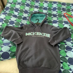 Svart hoodie från McKenzie med turkos huva - Snygg svart hoodie från McKenzie med stor logga framtill. Tröjan har turkos insida på huvan, snörning och en klassisk känguruficka. Perfekt för dig som gillar streetwear och vill ha en bekväm och stilren look.