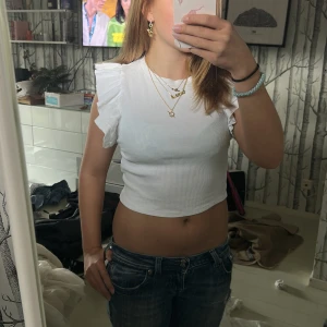 Vit croppad topp med volangärmar - Säljer en vit croppad topp med volangdetaljer på axlarna. Toppen är ribbad och har en rund halsringning samt ärmlös design med snygga volanger som ger en extra touch. Perfekt att matcha med jeans eller kjol för en cool look.