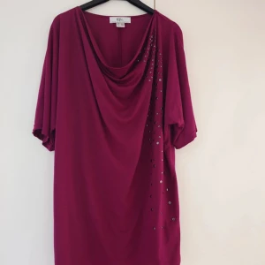 Vinröd kortklänning från b.p.c. - Snygg vinröd kortklänning från b.p.c. med draperad halsringning och dekorativa paljetter längs ena sidan. Klänningen har korta ärmar och är gjord i mjukt polyester-material som känns skönt mot huden. Perfekt för dig som gillar en chill och trendig look.