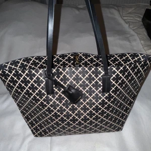 Malene Birger Abigail - Snygg shoppingväska i svart och beige med ett rutigt mönster. Väskan har långa svarta handtag och en praktisk dragkedja på insidan. Perfekt för att bära med sig allt man behöver under dagen.