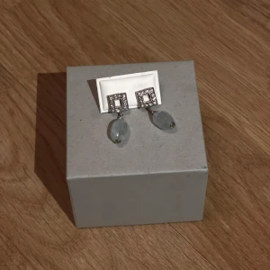 Örhängen med blå stenar och fyrkantig infattning - Eleganta örhängen med en fyrkantig infattning dekorerad med vita stenar och hängande blå stenar. Perfekta för att ge en touch av glamour till din outfit. Vet ej vad för material, köpte i Spanien i en smycke butik för ca 600 kr men har aldrig använt dessa. Det ser lite oxiderat ut, kanske silver?