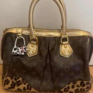 Brun handväska med leoparddetaljer  - Säljer en unik brun handväska från Louis Vuitton med klassiskt monogrammönster och leoparddetaljer i botten. Väskan har guldfärgade handtag och dragkedja samt en charmig hundformad nyckelring. Perfekt för att ge din outfit en lyxig touch.