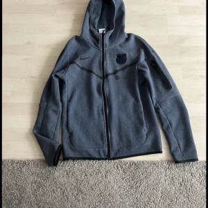 Navy blue Barca Nike tech - Säljer en grå huvtröja från Nike med dragkedja och klubbmärke på bröstet. Tröjan har långa ärmar och en praktisk ficka på ärmen. Perfekt för en sportig look eller avslappnade dagar.