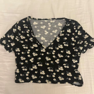 Blommig topp från Shein - Säljer en söt blommig topp från Shein i svart med vita och gula blommor. Toppen har en v-ringad design och korta ärmar. Den är croppad och har knytband i midjan för en justerbar passform. 