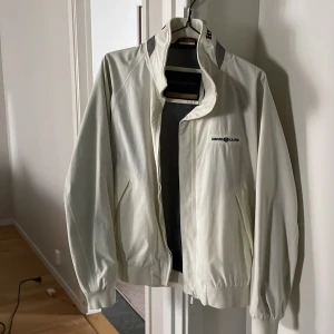 Vit vindjacka från Henri Lloyd - Säljer en stilren vit vindjacka från Henri Lloyd. Jackan har en dragkedja framtill och är långärmad med ribbade muddar. Perfekt för vår  och höst med sitt lätta och funktionella material. Färgen är cream