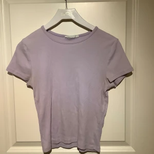 Ljuslila ribbad topp från KappAhl - Säljer en ljuslila ribbad topp från KappAhl. Toppen har en enkel design med korta ärmar och rund halsringning. Perfekt för en avslappnad stil.