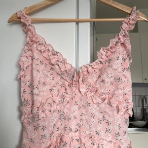 Rosa blommig klänning med volangdetaljer - Charmig rosa klänning med blommigt mönster och volangdetaljer. Perfekt för sommarens varma dagar. Klänningen har en v-ringad design och är ärmlös. 🩷