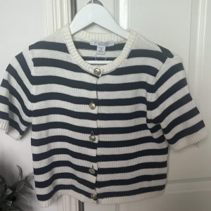 Randig kortärmad kofta - Säljer en snygg randig kofta i svart och vit. Den har korta ärmar och knappar framtill. Perfekt för en stilren look.