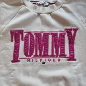 Vit sweatshirt från Tommy Hilfiger - Säljer en vit sweatshirt från Tommy Hilfiger med ett stort rosa logotryck på framsidan. Tröjan har långa ärmar och ribbade muddar vid ärmslut och nederkant. Perfekt för en avslappnad stil. Endast använd 2 gånger