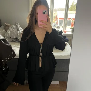 Svart blus med knytning - Snygg svart blus med knytning framtill och volangdetaljer. Blusen har långa ärmar och en v-ringad design som ger en elegant look. Perfekt för att klä upp en outfit!