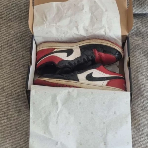 Nike Air Jordan 1 Low - Säljer ett par Nike Air Jordan 1 Low i klassisk svart, röd och vit färgkombination. Skorna har den ikoniska swoosh-loggan på sidan och en röd yttersula. Perfekta för sneakerheads som älskar en tidlös stil.