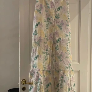 Blommig långkjol - Charmig långkjol med ett blommigt mönster i pastellfärger som lila, gul och grön. Klänningen har en luftig passform och är perfekt för varma dagar. Den har en volangdetalj som ger extra rörelse och stil.