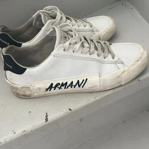 Vita sneakers från Armani Exchange - Snygga vita sneakers från Armani Exchange med svart text på sidan. Skorna har snörning och en rund tå. Perfekta för en stilren look. Men dom har ett hål i bakre insidan av skon men det syns inte när man har på skorna men annars är dom i mycket bra skick