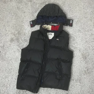 Svart dunväst från Tommy Hilfiger - Snygg svart dunväst från Tommy Hilfiger med avtagbar huva. Västen har dragkedja framtill och två praktiska fickor med dragkedjor. Insidan är fodrad i blått med en röd och vit detalj vid kragen. Storlek M Pris 800kr