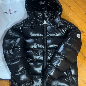 Moncler Maya - Snygg svart pufferjacka från Moncler med glansig finish och huva. Jackan har en dragkedja framtill och är perfekt för kyligare väder. Den ikoniska Moncler-loggan syns på ärmen. Dm för fler bilder! Tyvärr så är denna redan såld men har en exakt likadan, fast i storlek s/m. 
