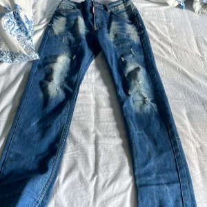 Blå jeans med slitningar från Dsquared2 - Snygga blå jeans från Dsquared2 med coola slitningar och en klassisk femficksdesign. De har en normal passform och är perfekta för en avslappnad stil. Jeansen har en dragkedja och knapp framtill. Har spänt dom inuti därför dom ser lite konstiga ut men annars jätte fint skick 