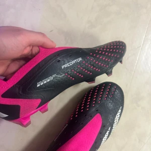Adidas Predator fotbollsskor - Snygga Adidas Predator fotbollsskor i svart och rosa med vita detaljer. Skorna har en modern design med en strömlinjeformad form och dobbar för optimalt grepp på planen. Perfekta för fotbollsspelare som vill ha både stil och funktion.