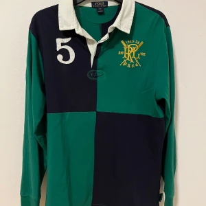 Långärmad pikétröja från Ralph Lauren - Säljer en snygg grön och marinblå pikétröja från Ralph Lauren med nummerdetalj och broderad logga på bröstet. Tröjan har en klassisk krage och långa ärmar, perfekt för en stilren look. Den är använd men i bra skick. Storlek 18y men passar som S-M