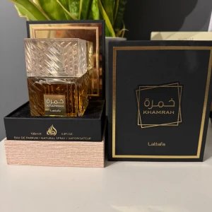 Khamrah Eau de Parfum från Lattafa - Elegant parfymflaska med ett lyxigt mönster och gyllene detaljer. Doften kommer i en fyrkantig glasflaska med en klar, geometrisk kork. Förpackningen är stilren med svart och guld design. Perfekt för den som söker en sofistikerad och unik doftupplevelse. 100ml kvar kom med bud!