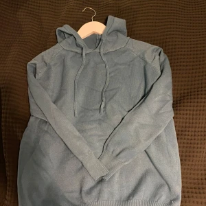 Blå merinoull hoodie - Säljer en snygg blå hoodie med långa ärmar och justerbar huva. Perfekt för en avslappnad stil. Den har ribbade muddar vid ärmslut och nederkant för extra komfort. Helt oanvänd, säljes pga har blivit för liten. Skulle säga att den passar 162-172