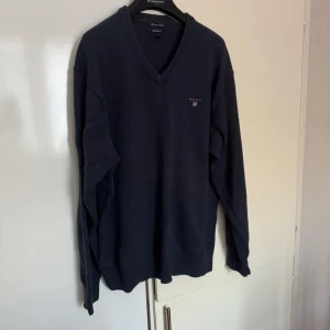 GANT - Säljer en stilren mörkblå tröja från GANT med v-ringning och broderad logga på bröstet. Tröjan har långa ärmar och ribbade kanter vid ärmslut och nederkant. Perfekt för en klassisk look.