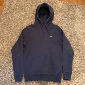 Mörkblå hoodie med lyle and scott logga - Säljer en stilren mörkblå hoodie med en liten gul logga på bröstet. Tröjan har en klassisk design med huva och känguruficka framtill. Perfekt för en avslappnad look.