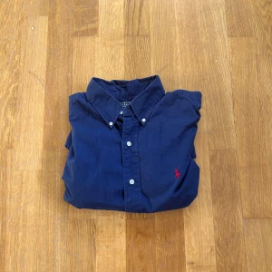 Mörkblå skjorta från Ralph Lauren - Säljer en klassisk mörkblå skjorta från Ralph Lauren med knappar och en broderad röd logotyp på bröstet. Skjortan har en stilren design med krage och är perfekt för en snygg och avslappnad look.