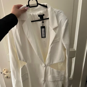 Vit kavaj från Boohoo - Snygg vit kavaj klänning från Boohoo med spetsdetaljer i midjan. Kavajen har en klassisk krage och knäpps med två knappar framtill. Perfekt för en stilren look. Prislappen kvar aldrig använd.