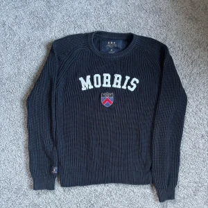Mörkblå stickad tröja från Morris - Säljer en snygg mörkblå stickad tröja från Morris med vit text och ett stilrent emblem på bröstet. Perfekt för en stilren look. Tröjan har långa ärmar och en rund halsringning.