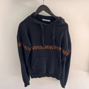Svart hoodie från NA-KD - Säljer en svart hoodie från NA-KD med en orange textdetalj runt bröstet. Den har en klassisk känguruficka och justerbar huva med dragsko. 