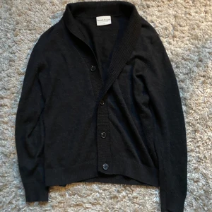 Cardigan Bruun & Stengade - Säljer en stilren svart kofta från Bruun & Stengade i merinoull. Koftan har en klassisk design med knappar framtill och ribbade kanter. Perfekt för att hålla sig varm under kyligare dagar. Skick: 8,5/10, Storlek L men sitter som M