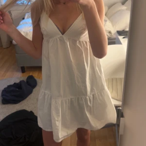 Vit ärmlös klänning - Säljer en söt vit klänning med smala axelband och volangdetaljer. Klänningen har en v-ringad design och knytning i ryggen. Perfekt för varma sommardagar!