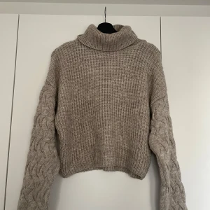 Beige stickad polotröja - Säljer en mysig beige stickad polotröja med flätmönstrade ärmar. Tröjan har en hög krage och är perfekt för kyliga dagar. Den är både stilren och bekväm, vilket gör den till ett måste i garderoben. Passar perfekt till jeans eller kjol för en avslappnad look.