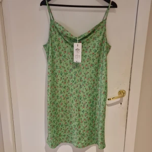 Grön blommig klänning med justerbara axelband - Söt grön klänning med blommigt mönster och justerbara axelband. Tillverkad i 100% polyester, perfekt för varma dagar. Klänningen har en avslappnad passform och ärmlös design.