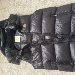 Svart dunväst från Moncler - Säljer en snygg svart dunväst från Moncler med dragkedja och huva. Perfekt för kyliga dagar när du vill hålla stilen. Västen har en quiltad design och är både praktisk och trendig.