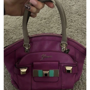 Lila handväska från Guess - Snygg lila handväska från Guess med gulddetaljer. Väskan har ett färgglatt blommigt foder och en praktisk dragkedja upptill. Perfekt för att ge en färgklick till din outfit!