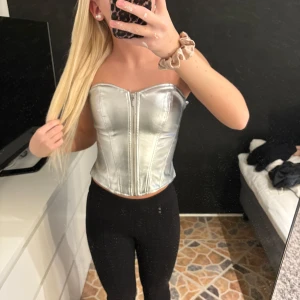 Silverfärgad korsett - Snygg och glansig silverfärgad korsett med en axelbandslös design. Korsetten har en tight passform som framhäver figuren. Aldrig använd!