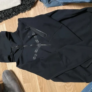 Svart Nike tech  - Säljer en svart jacka från Nike med dragkedja och huva. Jackan har en diskret logga på bröstet och en ficka med dragkedja på ärmen. Perfekt för en sportig look!