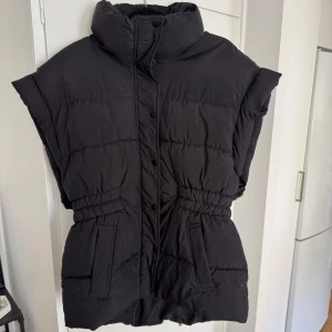 Svart dunväst med dragkedja - Snygg svart dunväst med hög krage och dragkedja framtill. Västen har en elastisk midja som gen en figursydd passform, fickor på sidorna. Sprickigt lite i ”armhålan” på båda sidor som ni ser på sista bilden, men inget man ser, samt går lätt att sy om man skulle vilja.