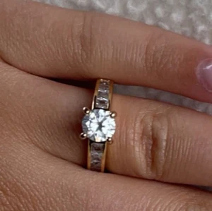 Guldfärgad ring med stor diamand - Elegant guldfärgad ring med en stor, glittrande diamand i mitten och mindre diamanter på sidorna. Perfekt för att ge en touch av glamour till din stil. 