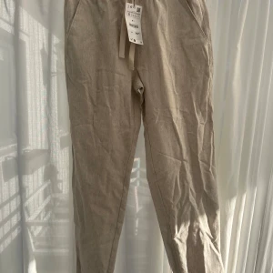 Beige linnebyxor från Zara - Snygga beige linnebyxor från Zara med elastisk midja och snörning. Perfekta för en avslappnad stil. Byxorna har en slim fit och är tillverkade i ett lätt och luftigt material.