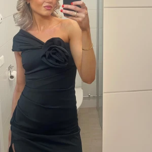 Slutsåld klänning från ASOS - Elegant svart oneshoulder klänning med en stor rosdetalj på axeln och slits. Storlek 36. Slutsåld på ASOS. Ord pris 1349kr.
