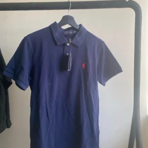Blå pikétröja från Ralph Lauren - Säljer en klassisk blå pikétröja från Ralph Lauren med korta ärmar och den ikoniska röda loggan på bröstet. Perfekt för en stilren och avslappnad look. Tröjan har en krage och knappar framtill. Passar utmärkt till både jeans och chinos.